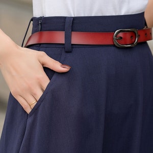 May include: Close-up of a navy blue skirt with a red leather belt. The belt has a dark metal buckle. A hand is in the skirt pocket, and a gold ring is visible. The skirt appears to be made of a lightweight fabric.