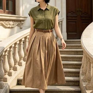 May include: A woman's outfit featuring a short-sleeved olive green button-down shirt, a high-waisted pleated tan skirt, and a brown belt. The outfit is accessorized with a brown hat and black sandals. The woman is walking down a stone staircase.