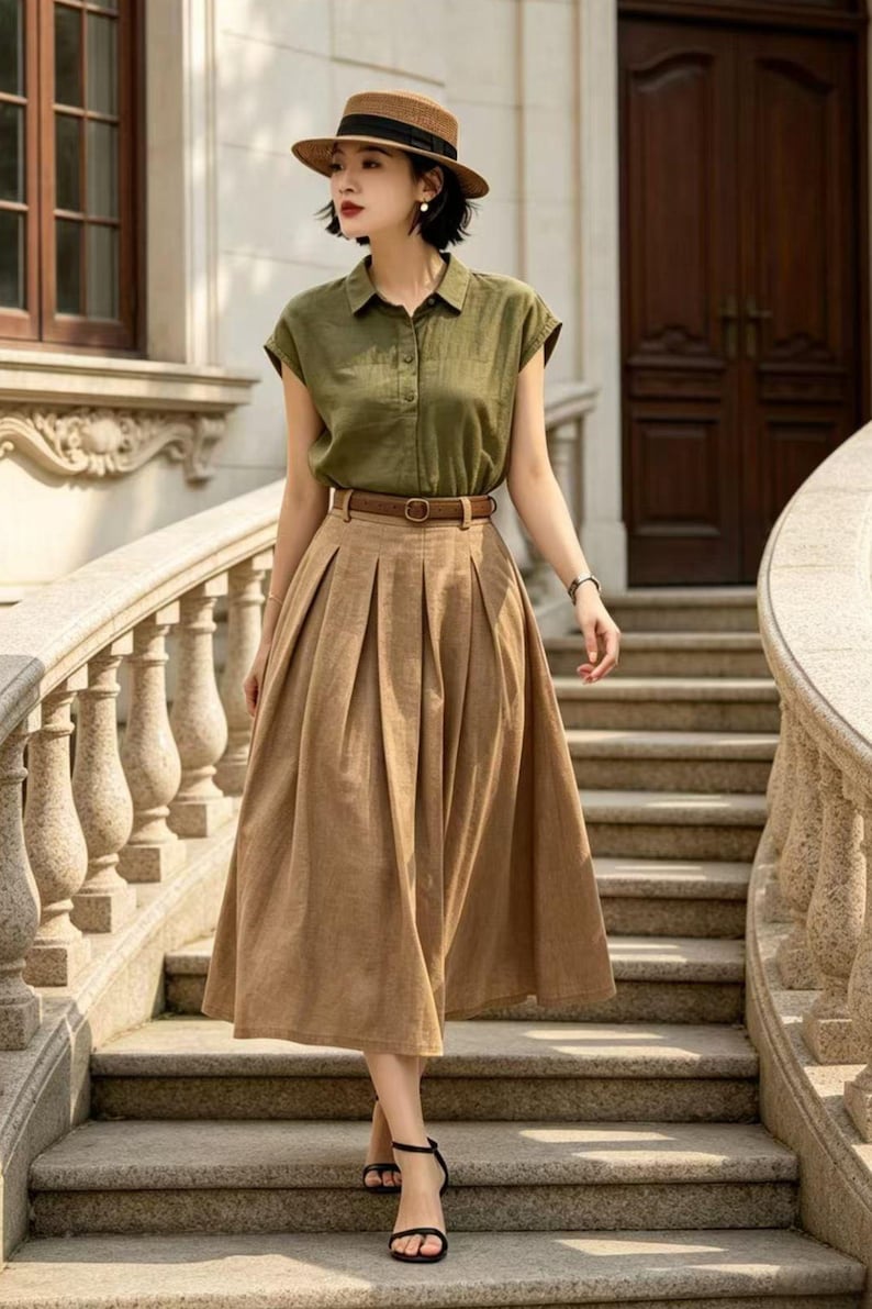 May include: A woman's outfit featuring a short-sleeved olive green button-down shirt, a high-waisted pleated tan skirt, and a brown belt. The outfit is accessorized with a brown hat and black sandals. The woman is walking down a stone staircase.