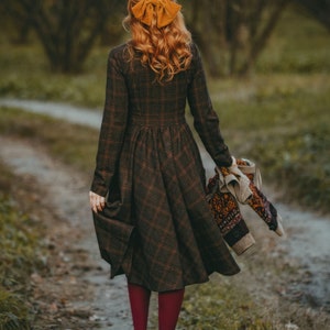 Pu&ograve; includere: Una donna con un vestito a quadri marroni e neri e un fiocco giallo nei capelli cammina lungo un sentiero in un bosco. Tiene in mano un maglione di lana.