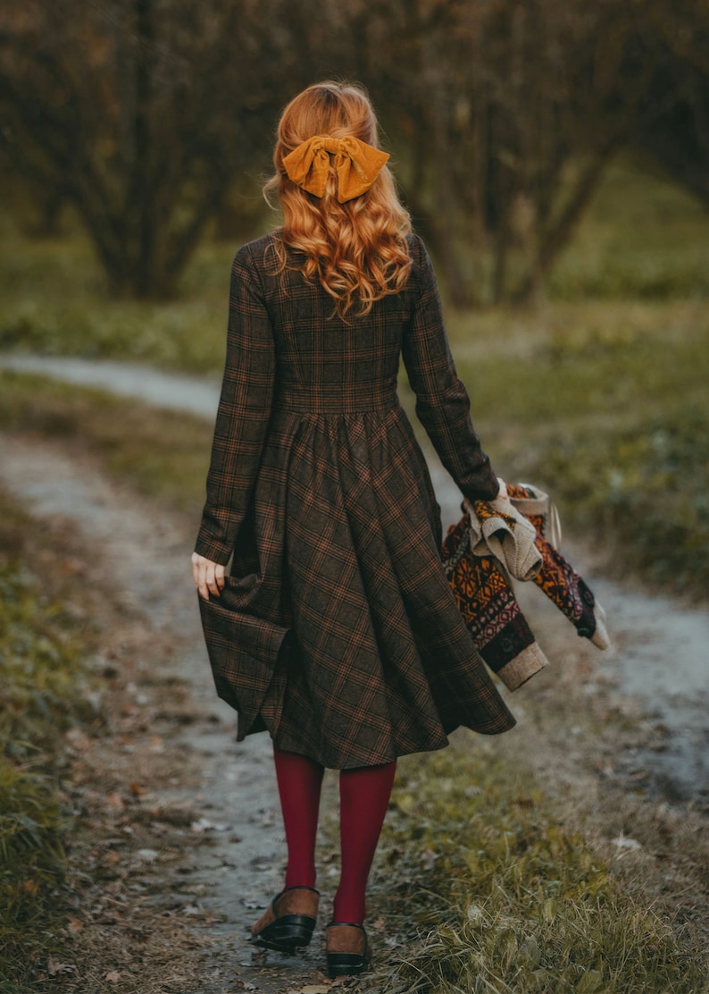 Pu&ograve; includere: Una donna con un vestito a quadri marroni e neri e un fiocco giallo nei capelli cammina lungo un sentiero in un bosco. Tiene in mano un maglione di lana.