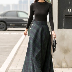 May include: A woman wearing a black turtleneck sweater and a green and black plaid skirt. She is wearing a black hat with white lettering and black boots. She is carrying a black handbag.