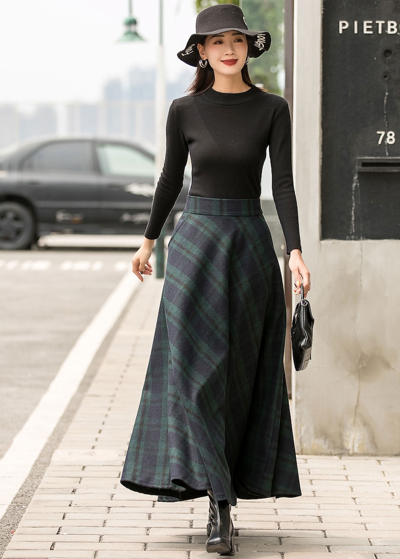 May include: A woman wearing a black turtleneck sweater and a green and black plaid skirt. She is wearing a black hat with white lettering and black boots. She is carrying a black handbag.