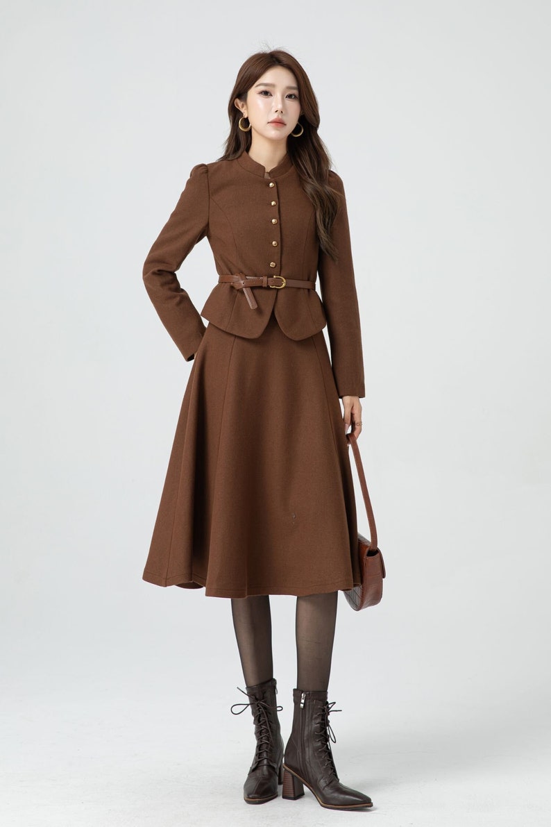 May include: A brown skirt suit with a fitted jacket and a flared skirt. The jacket has a mandarin collar and gold buttons. The skirt has a brown leather belt with a gold buckle. The model is wearing brown lace-up boots.