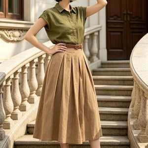 May include: A woman wearing a short-sleeved olive green button-down shirt, a brown pleated midi skirt, and a brown belt. She is wearing a brown hat and black sandals. The outfit is suitable for warm weather.