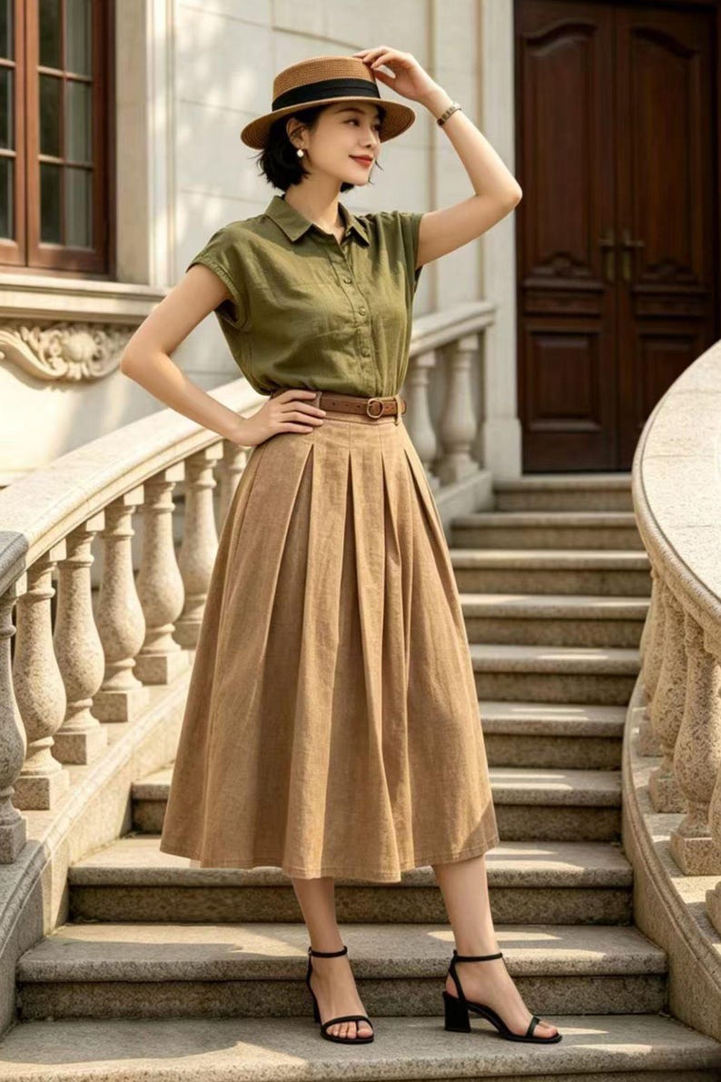 May include: A woman wearing a short-sleeved olive green button-down shirt, a brown pleated midi skirt, and a brown belt. She is wearing a brown hat and black sandals. The outfit is suitable for warm weather.