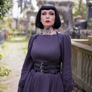 May include: A woman with black hair and tattoos is wearing a dark purple dress with a black leather harness and a wide belt with silver buckles. She is standing in a cemetery.