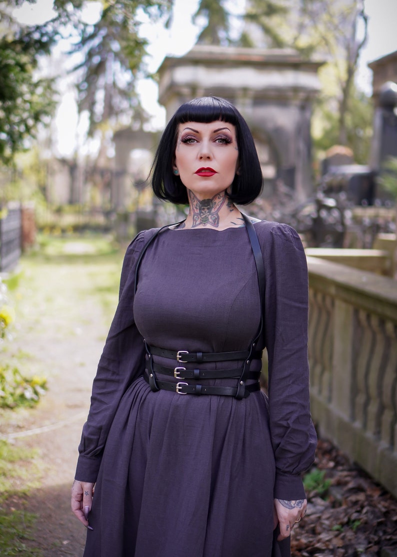 May include: A woman with black hair and tattoos is wearing a dark purple dress with a black leather harness and a wide belt with silver buckles. She is standing in a cemetery.