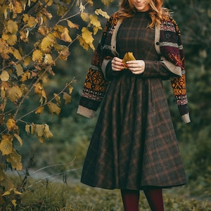 Pu&ograve; includere: Una donna indossa un vestito a quadri marroni e neri con un cardigan marrone con motivi geometrici colorati. Tiene una foglia gialla nelle mani. Il vestito ha una gonna ampia e il cardigan ha maniche lunghe.