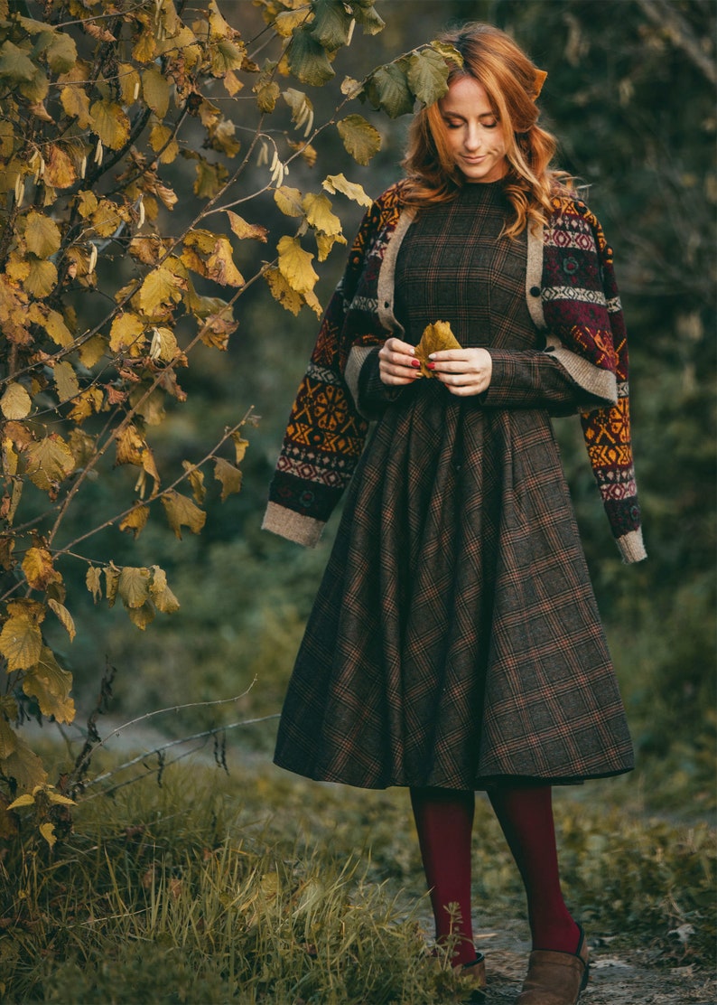 Pu&ograve; includere: Una donna indossa un vestito a quadri marroni e neri con un cardigan marrone con motivi geometrici colorati. Tiene una foglia gialla nelle mani. Il vestito ha una gonna ampia e il cardigan ha maniche lunghe.