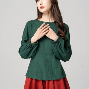 May include: A green long-sleeved blouse with a round neckline and gathered sleeves. The blouse is tucked into a red skirt.