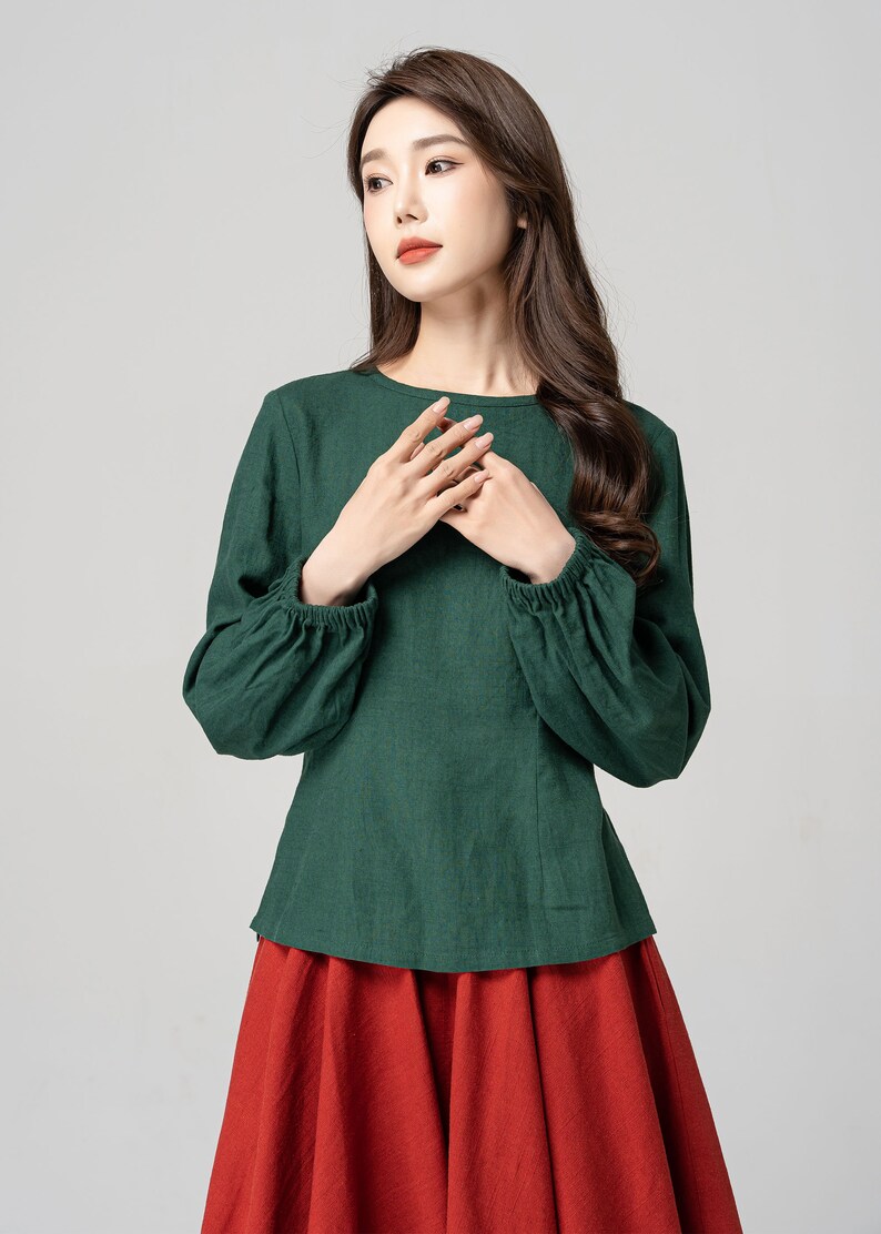 May include: A green long-sleeved blouse with a round neckline and gathered sleeves. The blouse is tucked into a red skirt.
