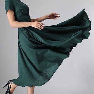 Puede incluir: Un vestido midi de color verde oscuro con mangas cortas, cuello redondo y una falda amplia y acampanada que se extiende hacia afuera. La prenda presenta un acabado de tela texturizada y se combina con zapatos de tac&oacute;n alto negros con plataforma.