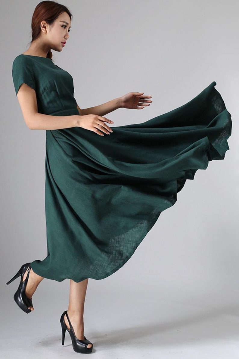 Puede incluir: Un vestido midi de color verde oscuro con mangas cortas, cuello redondo y una falda amplia y acampanada que se extiende hacia afuera. La prenda presenta un acabado de tela texturizada y se combina con zapatos de tac&oacute;n alto negros con plataforma.