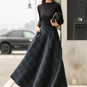 May include: A woman wearing a black long-sleeved shirt and a black hat with white lettering. She is wearing a green and black plaid skirt and black heeled boots. She is holding a black purse.