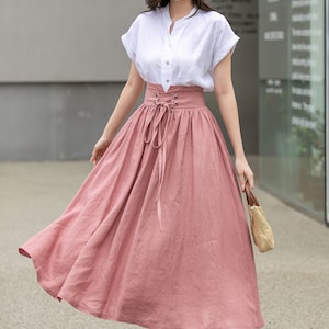 May include: A woman wearing a white linen shirt with a pink linen skirt. The skirt has a lace-up detail at the waist and a flowing silhouette.