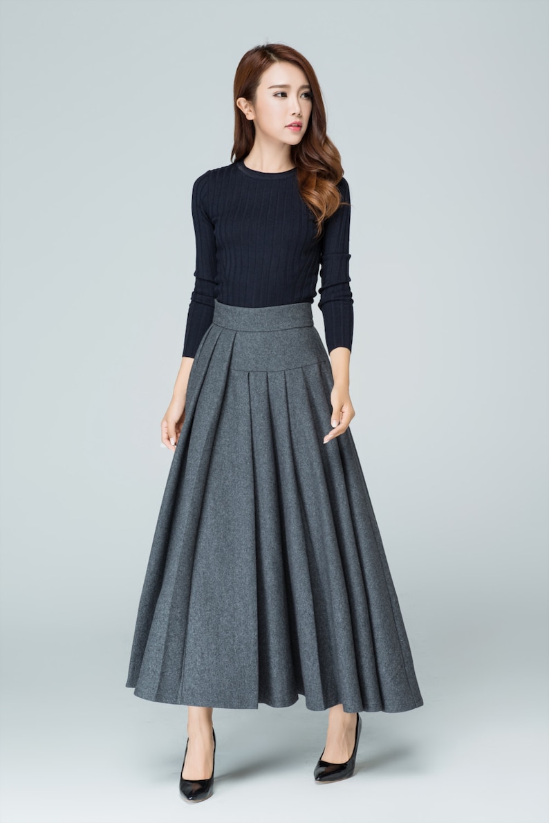 May include: A woman wearing a black long-sleeved sweater and a gray pleated maxi skirt. She is standing on a white background.