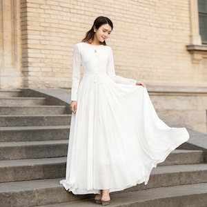 Puede incluir: Un vestido largo y blanco con mangas largas y escote de ojo de cerradura. El vestido presenta una cintura ce&ntilde;ida con un lazo y una falda larga hasta el suelo. La modelo est&aacute; de pie sobre unos escalones de piedra.