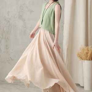 May include: A woman wearing a light green sleeveless top and a flowing, pale pink skirt. The skirt has a ruffled hem and appears to be made of a lightweight fabric. The outfit is paired with a black necklace.
