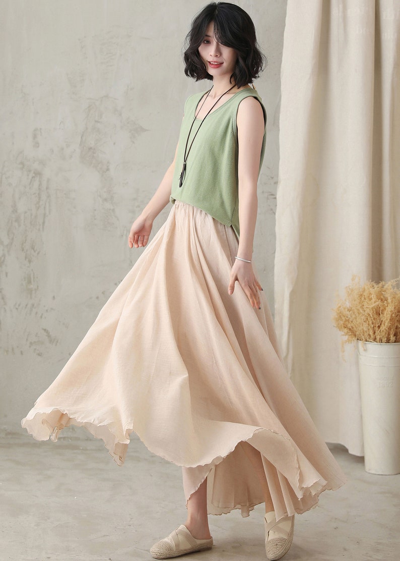 May include: A woman wearing a light green sleeveless top and a flowing, pale pink skirt. The skirt has a ruffled hem and appears to be made of a lightweight fabric. The outfit is paired with a black necklace.