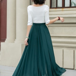 May include: A woman wearing a white top and a long, flowing emerald green skirt. The skirt is gathered at the waist and has a flowing, A-line silhouette.