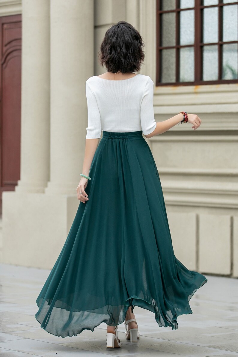 May include: A woman wearing a white top and a long, flowing emerald green skirt. The skirt is gathered at the waist and has a flowing, A-line silhouette.