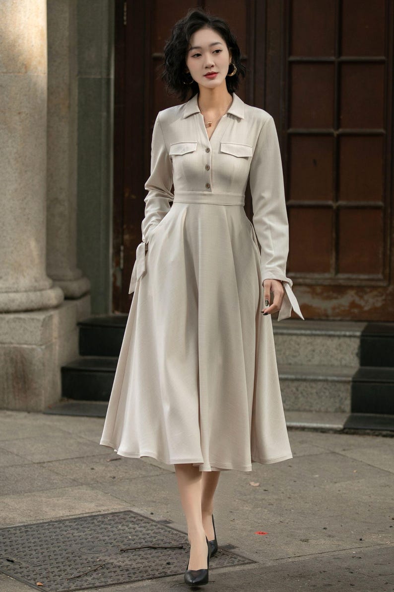 May include: A beige long-sleeved dress with a collared neckline, button closure, and two pockets. The dress has a fitted bodice and a flowing skirt.