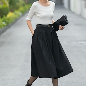 May include: A woman wearing a white, short-sleeved top, a black pleated skirt, and black ankle boots. She is holding a black clutch and a black belt with a gold buckle. The outfit is suitable for business or casual wear.