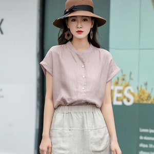 May include: A woman wearing a light pink linen blouse with short sleeves and a button-up front. She is also wearing a beige linen skirt with pockets and a brown straw hat with a black ribbon.