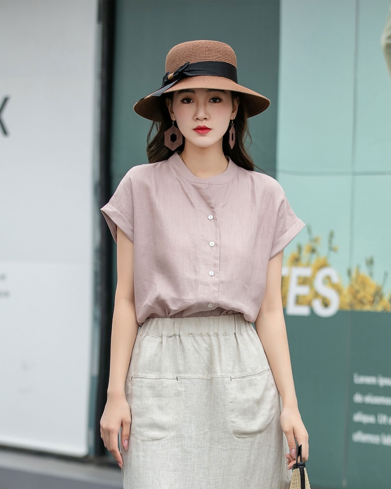May include: A woman wearing a light pink linen blouse with short sleeves and a button-up front. She is also wearing a beige linen skirt with pockets and a brown straw hat with a black ribbon.