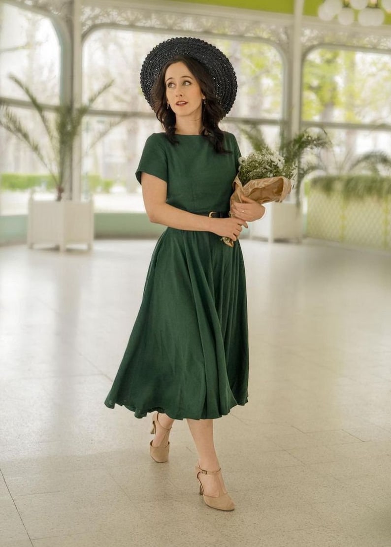 May include: A woman wearing a green midi dress with a black hat and holding a bouquet of white flowers.