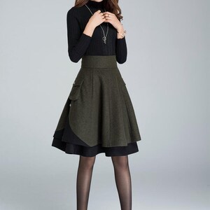 Puede incluir: Una mujer con un jersey negro de cuello alto y una falda de capas verde y negra. La falda tiene cintura alta y silueta acampanada. Lleva medias negras y tacones altos negros.