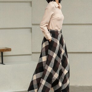 May include: A woman wearing a beige plaid button-down shirt and a long, brown, black, and cream plaid skirt. She is wearing dark brown lace-up boots. The outfit is a classic, stylish ensemble.