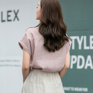 May include: A woman wearing a light pink linen shirt with short sleeves and a light beige linen skirt. The shirt has a collar and a button-up front. The skirt has a high waist and a flowy silhouette.