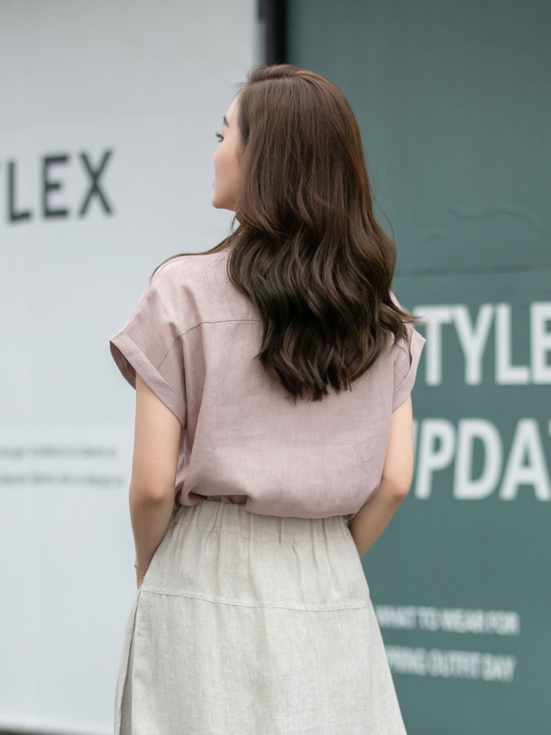 May include: A woman wearing a light pink linen shirt with short sleeves and a light beige linen skirt. The shirt has a collar and a button-up front. The skirt has a high waist and a flowy silhouette.