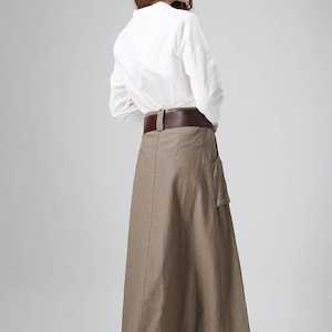 May include: A long brown linen skirt with a brown leather belt and a large pocket on the right side. The skirt is worn with a white button-down shirt.