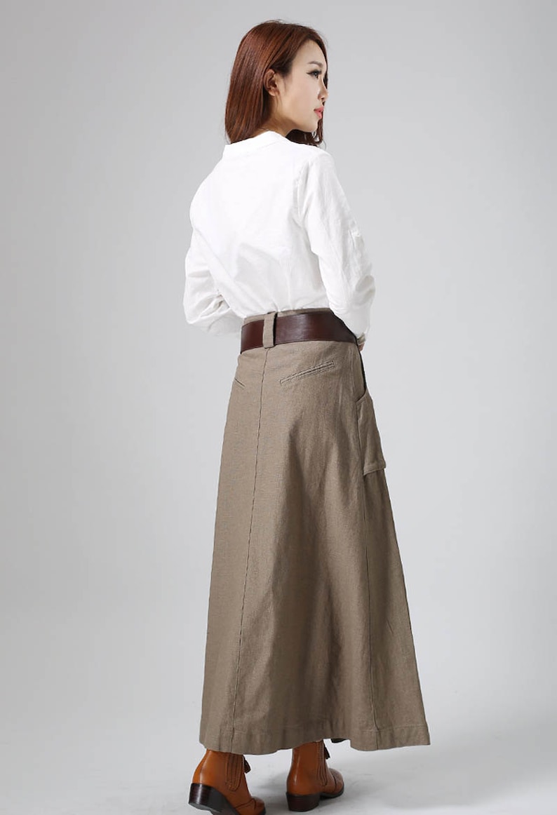 May include: A long brown linen skirt with a brown leather belt and a large pocket on the right side. The skirt is worn with a white button-down shirt.