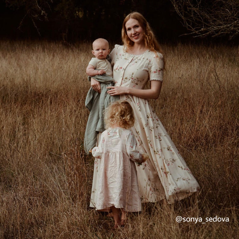 May include: A woman in a floral dress holds a baby in her arms while a young girl in a pink dress stands in front of her. The family is standing in a field of tall grass.