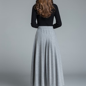 May include: A long, gray skirt with a pleated waist and a black long-sleeved top. The skirt is a solid color and falls to the ankles. The model is wearing black boots. The outfit is suitable for fall or winter.
