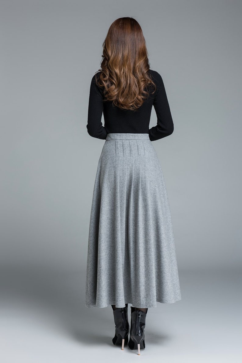 May include: A long, gray skirt with a pleated waist and a black long-sleeved top. The skirt is a solid color and falls to the ankles. The model is wearing black boots. The outfit is suitable for fall or winter.