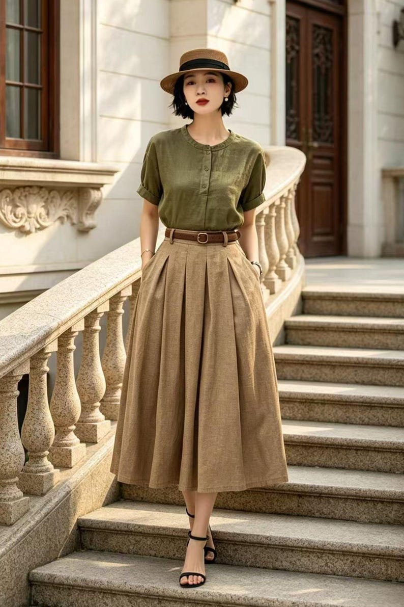 May include: A woman's outfit featuring a short-sleeved olive green button-up shirt, a brown belt, and a long, pleated tan skirt. The outfit is completed with a brown hat and black sandals. The ensemble is suitable for a casual or semi-formal occasion.