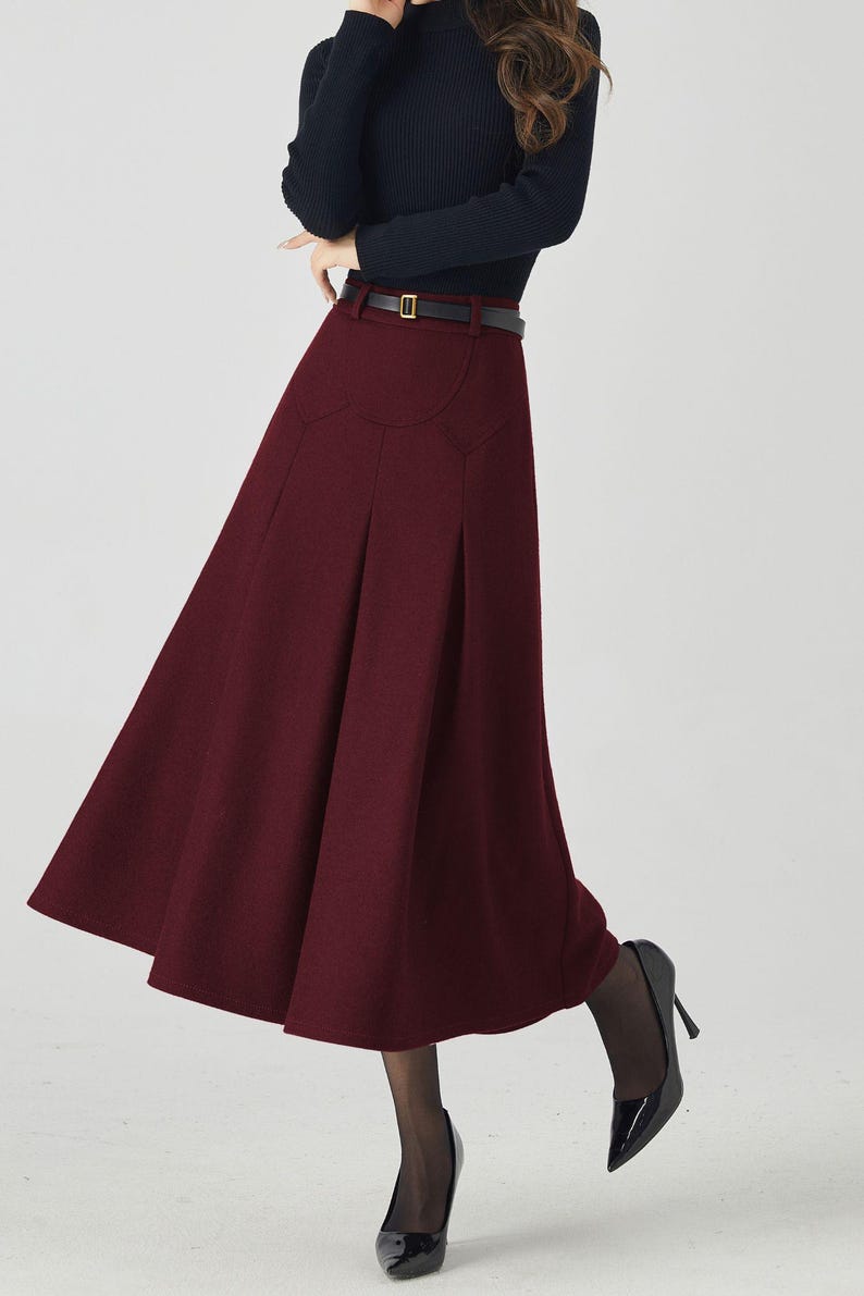 May include: A burgundy midi skirt with a high waist and a flared silhouette. The skirt is paired with a black long-sleeve top and a black belt. Black high heels complete the outfit.