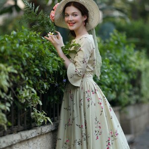 Peut inclure: Une femme porte une robe cr&egrave;me &agrave; motifs floraux et un chapeau de paille &agrave; larges bords orn&eacute; de fleurs roses. La robe a des manches longues et une taille ajust&eacute;e. Elle sourit pr&egrave;s d'une haie verte.