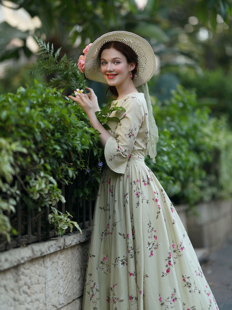 Peut inclure: Une femme porte une robe cr&egrave;me &agrave; motifs floraux et un chapeau de paille &agrave; larges bords orn&eacute; de fleurs roses. La robe a des manches longues et une taille ajust&eacute;e. Elle sourit pr&egrave;s d'une haie verte.