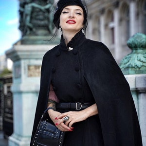 May include: A woman wearing a black cape with a black hat and a black handbag. She is wearing a black belt with a silver buckle. She is standing in front of a stone building.