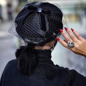 May include: A black velvet beret with a black net veil and a black bow. The beret has a small black button on the bow. The person wearing the beret has red nail polish and is wearing a black ring with a skull design.