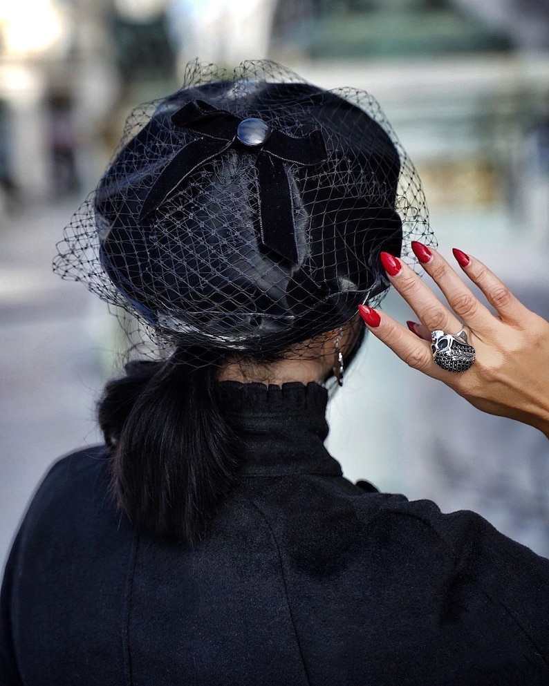 May include: A black velvet beret with a black net veil and a black bow. The beret has a small black button on the bow. The person wearing the beret has red nail polish and is wearing a black ring with a skull design.