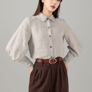 Può includere: Una camicia grigio chiaro con bottoni, a righe verticali e maniche lunghe. Abbinata a pantaloni marroni a pieghe e una cintura marrone con fibbia dorata. L'outfit è uno stile classico ed elegante.