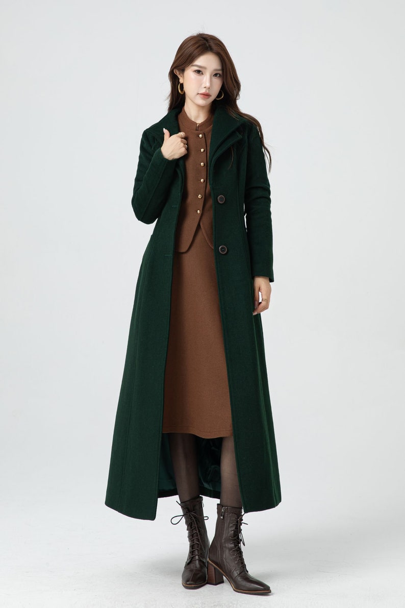 May include: A long, dark green coat with a high collar and button closure. The coat is worn over a brown dress and brown boots.