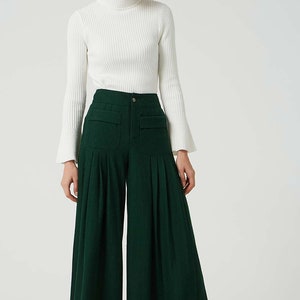 May include: A woman is wearing a white turtleneck sweater and dark green wide-leg trousers with pleats. The trousers have large pockets on the front.
